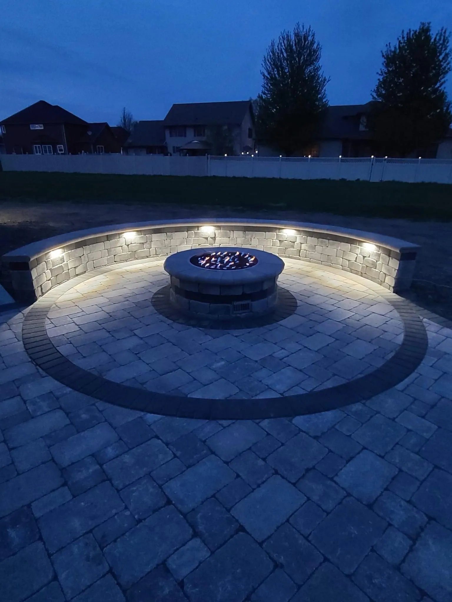 Circular paver patio with a curved seat wall, built-in lighting, and a round gas fire pit at dusk in Chicago's Southwest Suburbs