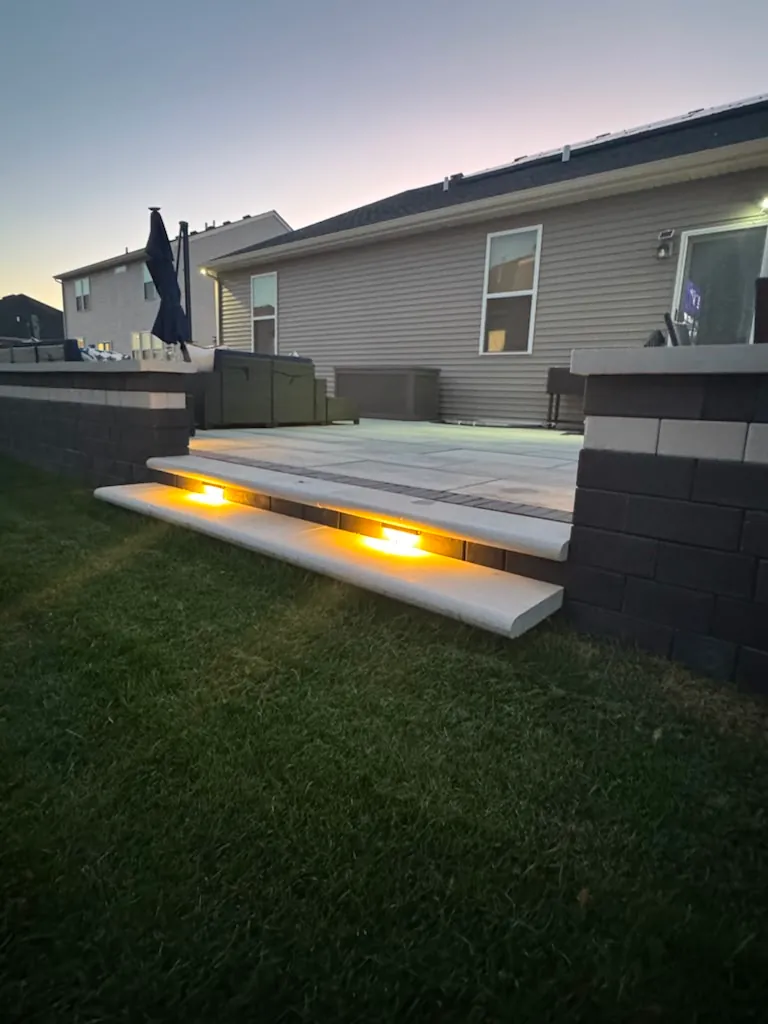 Raised large-format paver patio with wide cap-stone steps, recessed step lighting, and two-tone retaining and seat walls at dusk in Chicago's Southwest Suburbs